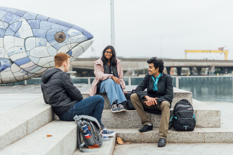 Three students sit beside big fish in front of the Lagan
