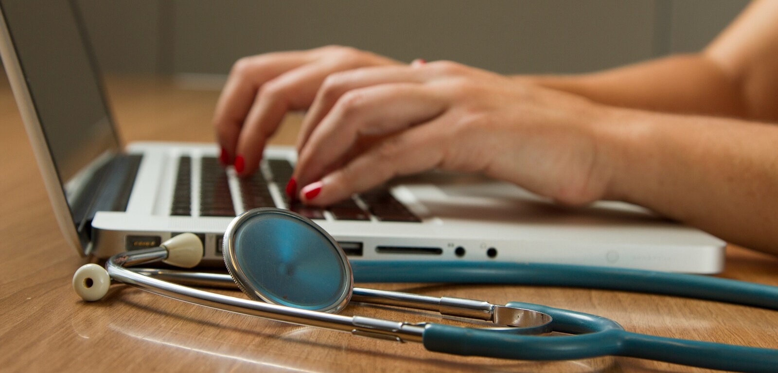 Hands typing on a laptop with a stethoscope nearby