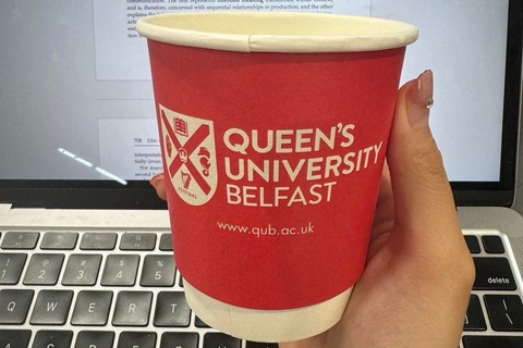 Student holds paper QUB coffee cup in front of laptop