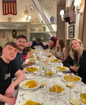A group of smiling students seated at a long table ina restaurant with dishes of paella in front of them