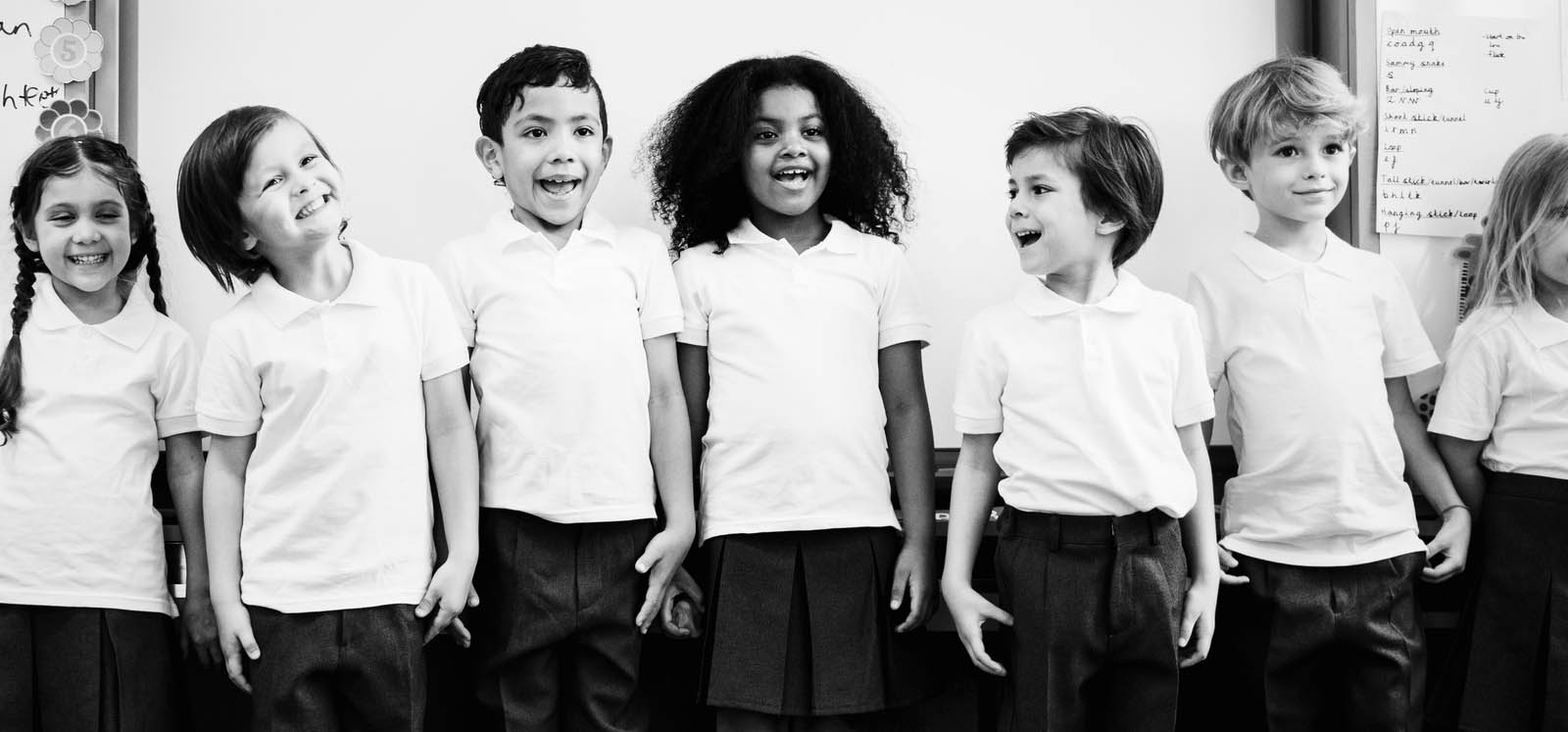 A group of six young school children standing in a row, smiling and laughing, dressed in school uniforms with white polo shirts and dark trousers or skirts, in a classroom setting.